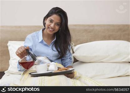 Portrait of beautiful young woman pouring tea in cup while sitting in bed