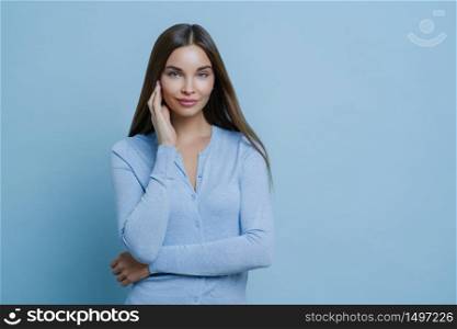 Portrait of beautiful young woman touches face gently, wears blue jumper, has appealing appearance, poses indoor against blue background, knows how to attract mens attention, flirts over camera