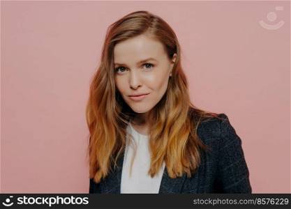 Portrait of beautiful young woman with ginger hair looking at camera confidently, wearing dark formal jacket and white top posing isolated against pink studio wall. Human emotions concept. Attractive young woman with ginger hair in formal jacket posing against pink background