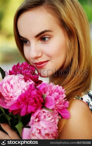 Portrait of beautiful young woman with long blonde hair holding bouquet of pink peonies. Fashion model girl with bunch of flowers in park in summer.