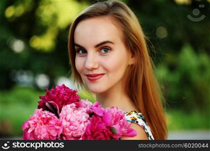 Portrait of beautiful young woman with long blonde hair holding bouquet of pink peonies. Fashion model girl with bunch of flowers in park in summer.