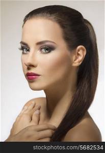 Portrait of beautiful, youth woman on grey background. She has long, straight, brown hair clipped in ponytail, strong purple make up and she is loking at the camera.