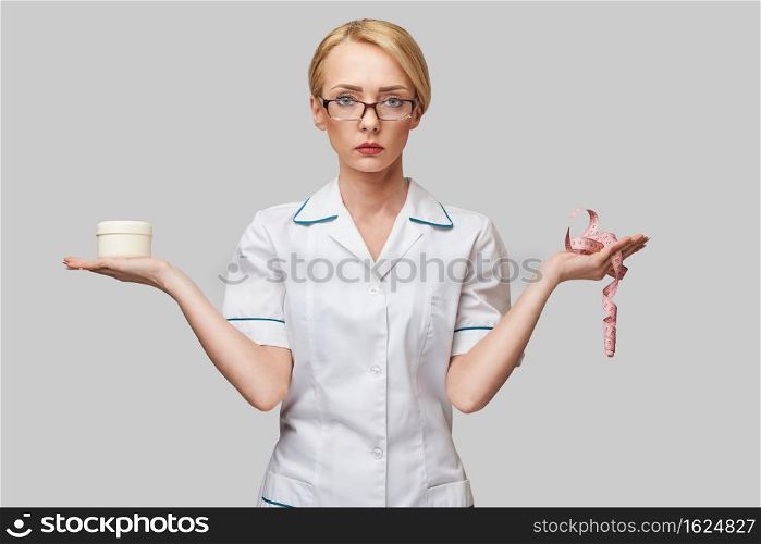 Portrait of beauty specialist standing against grey background and holding in her hand a face cream jar and measure tape.. Portrait of beauty specialist standing against grey background and holding in her hand a face cream jar and measure tape