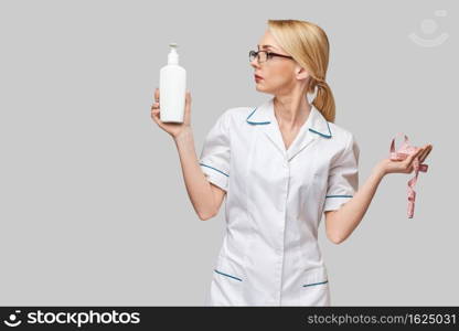 Portrait of beauty specialist standing against grey background and holding in her hand bottle of body lotion and measure tape.. Portrait of beauty specialist standing against grey background and holding in her hand bottle of body lotion and measure tape