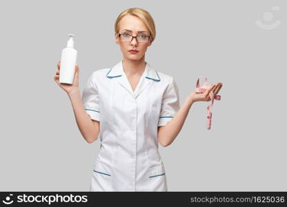 Portrait of beauty specialist standing against grey background and holding in her hand bottle of body lotion and measure tape.. Portrait of beauty specialist standing against grey background and holding in her hand bottle of body lotion and measure tape