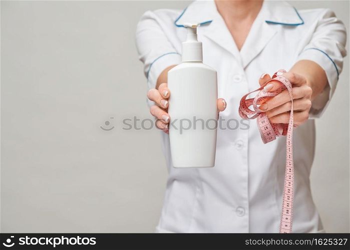 Portrait of beauty specialist standing against grey background and holding in her hand bottle of body lotion and measure tape.. Portrait of beauty specialist standing against grey background and holding in her hand bottle of body lotion and measure tape
