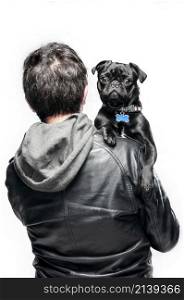 portrait of black pug and man, the pug is looking at the camera in the arms of the owner who is not facing the camera, the owner has a black hoodie.