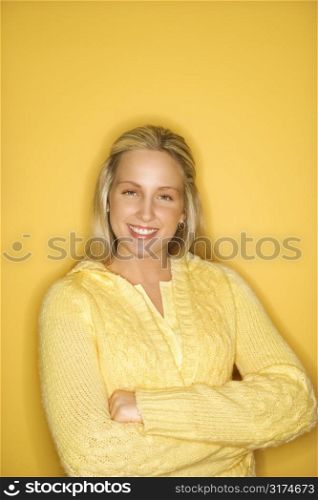 Portrait of blond Caucasian teen girl smiling with crossed arms against yellow background.