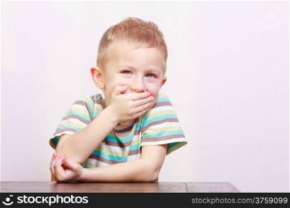 Portrait of blond happy laughing boy child kid covering mouth with his hand at the table interior. Emotions and fun.