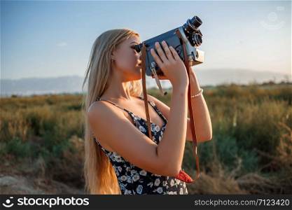 portrait of blonde girl in floral print dress with vintage video camera in grape field during sunset
