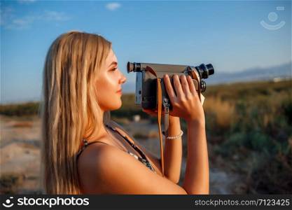 portrait of blonde girl in floral print dress with vintage video camera in grape field during sunset