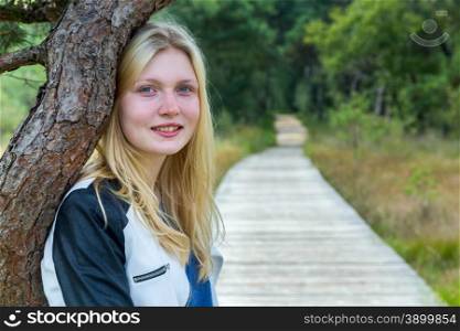Portrait of blonde teenage girl with trunk and wooden footpath in forest nature