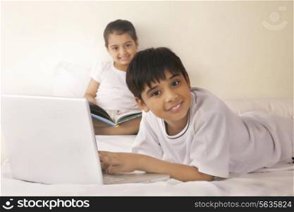 Portrait of boy using laptop with girl reading book in bed