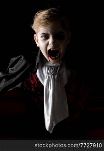 Portrait of boy wearing Halloween vampire makeup and costume cloak bare his teeth on black background.. Boy in Halloween vampire makeup costume