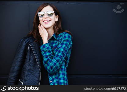 Portrait of brunette girl wearing leather jacket standing outdoors in the city against the black urban wall. Brunette girl in rock black style, wearing leather jacket standing outdoors in the city against the black urban wall.