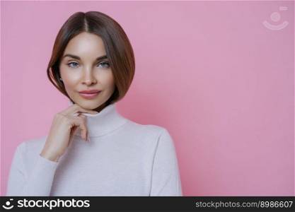 Portrait of brunette woman keeps hand on collar of turtleneck, has healthy perfect skin, wears minimal makeup, has serious expression, poses against pink background. Human facial expressions concept