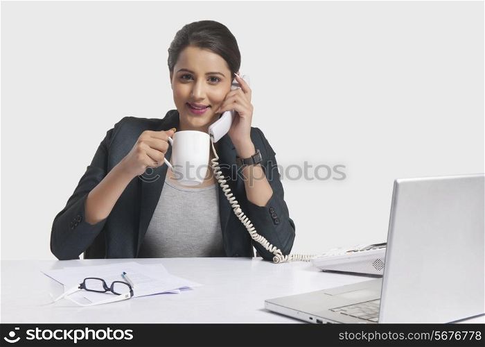 Portrait of businesswoman having coffee while answering telephone at desk against white background
