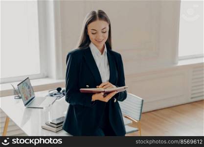 Portrait of busy business woman with dark brown straight hair in black formal suit writing down and looking at note book, leaning on sideways desk in her cabinet with two windows on background. Focused business woman writing down business plan in notebook while leaning on desk