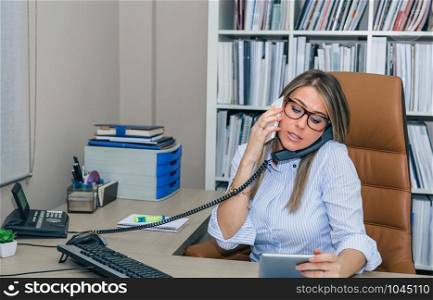 Portrait of busy businesswoman talking simultaniously on cell and stationary phone while looking electronic tablet in her hands. Busy businesswoman talking with cell and fixed line phone in office