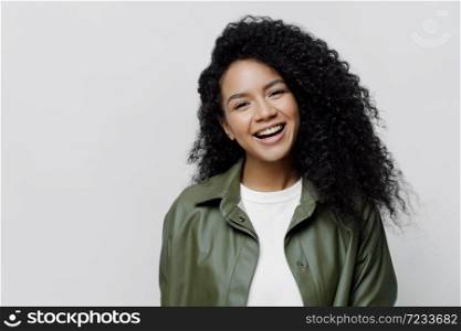 Portrait of carefree joyful African American lady with beaming smile and curly bushy hair, glad to be promoted at work, hears excellent news, dressed in leather shirt, isolated on white background