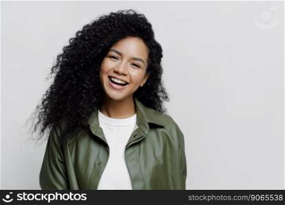 Portrait of carefree joyful African American lady with beaming smile and curly bushy hair, glad to be promoted at work, hears excellent news, dressed in leather shirt, isolated on white background