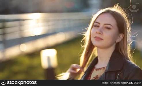 Portrait of caucaisian girl with long blonde straight hair and magic brown eyes looking at camera with shy smile in glow of amazing sunset. Beautiful young female with clean skin smiling outdoors. Positive emotion and facial expression.