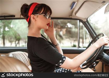 portrait of Caucasian beautiful young girl in black vintage dress and vintage glasses posing behind the wheel of a vintage car