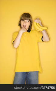 Portrait of Caucasian boy holding bunch of bananas and gesturing with finger in mouth that they are gross standing against yellow background.