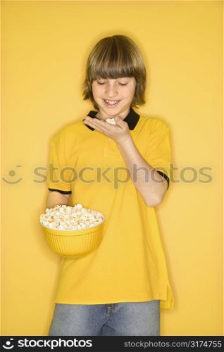 Portrait of Caucasian boy smiling holding bowl of popcorn in one hand and bringing some popcorn to mouth standing against yellow background.