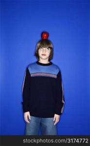 Portrait of Caucasian boy with an apple on his head standing against blue background.