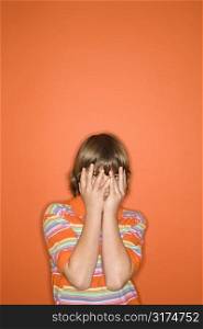 Portrait of Caucasian boy with hands covering his face standing against orange background.