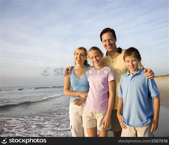 Portrait of Caucasian family of four posing on beach looking at viewer smiling.