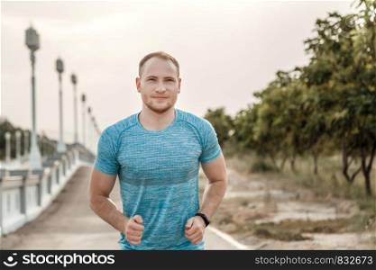 portrait of Caucasian guy in a blue t-shirt and black shorts who trains and runs on the asphalt track during sunset