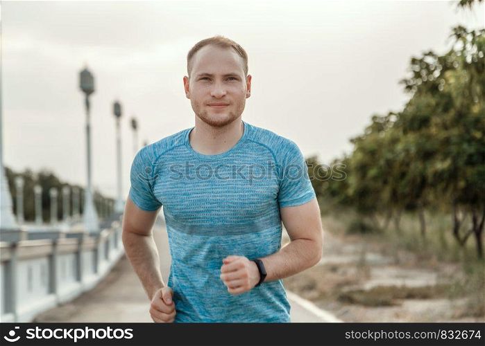 portrait of Caucasian guy in a blue t-shirt and black shorts who trains and runs on the asphalt track during sunset