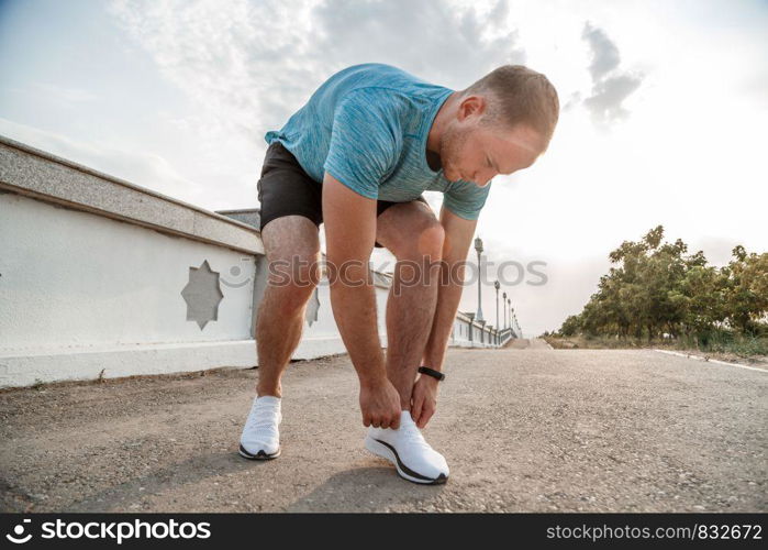 portrait of Caucasian guy in a blue t-shirt and black shorts,who ties his laces on white sports shoes before Jogging