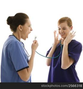 Portrait of Caucasian healthcare workers wearing scrubs with one yelling into stethoscope that is attached to other&acute;s ears against white background.