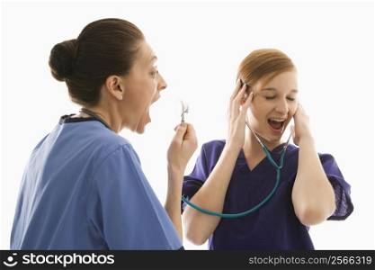Portrait of Caucasian healthcare workers wearing scrubs with one yelling into stethoscope that is attached to other&acute;s ears against white background.