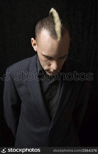 Portrait of Caucasian man in suit with mohawk looking down against black background.