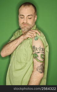 Portrait of Caucasian man pulling up sleeve to show tattoo standing against green background.