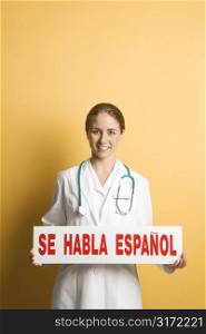 Portrait of Caucasian mid-adult female doctor holding up se habla espanol sign against yellow background smiling and looking at viewer.