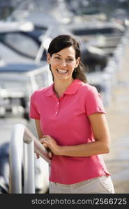 Portrait of Caucasian mid-adult female smiling at camera and holding railing at harbor.
