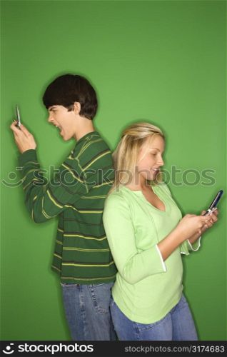 Portrait of Caucasian teen boy and girl on cellphones standing with backs to eachother against green background.