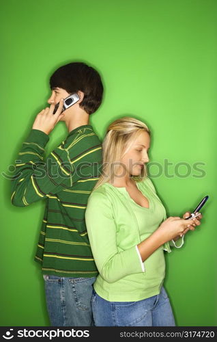 Portrait of Caucasian teen boy and girl on cellphones standing with backs to eachother against green background.