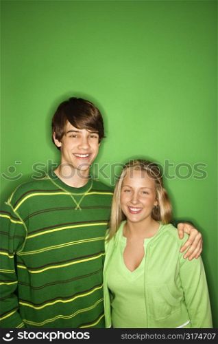 Portrait of Caucasian teen boy and girl standing against green background smiling with arms around eachother.