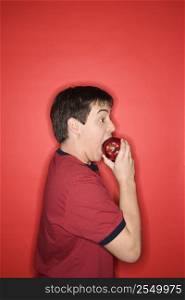 Portrait of Caucasian teen boy biting an apple standing against red background.