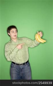 Portrait of Caucasian teen boy holding and pointing at bananas standing against green background.