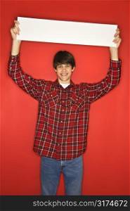 Portrait of Caucasian teen boy holding blank sign above his head standing against red background.