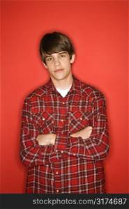 Portrait of Caucasian teen boy with arms crossed standing against red background wearing flannel shirt.