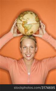 Portrait of Caucasian teen girl holding globe on her head smiling standing against orange background.