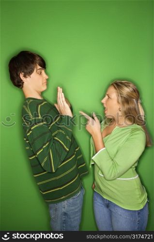 Portrait of Caucasian teen girl pointing at yelling at teen boy who is holding up hands to her against green background.